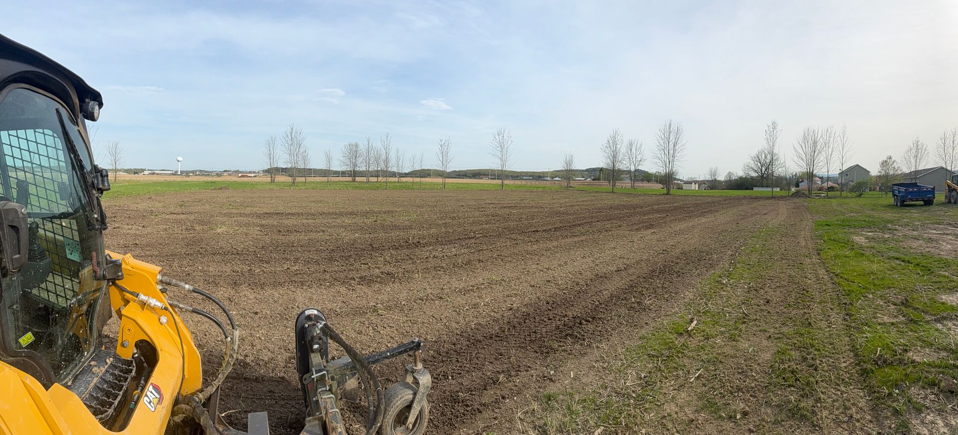 A yellow tractor tilling a field under a cloudy sky. Bare trees and power lines are visible in the distance.