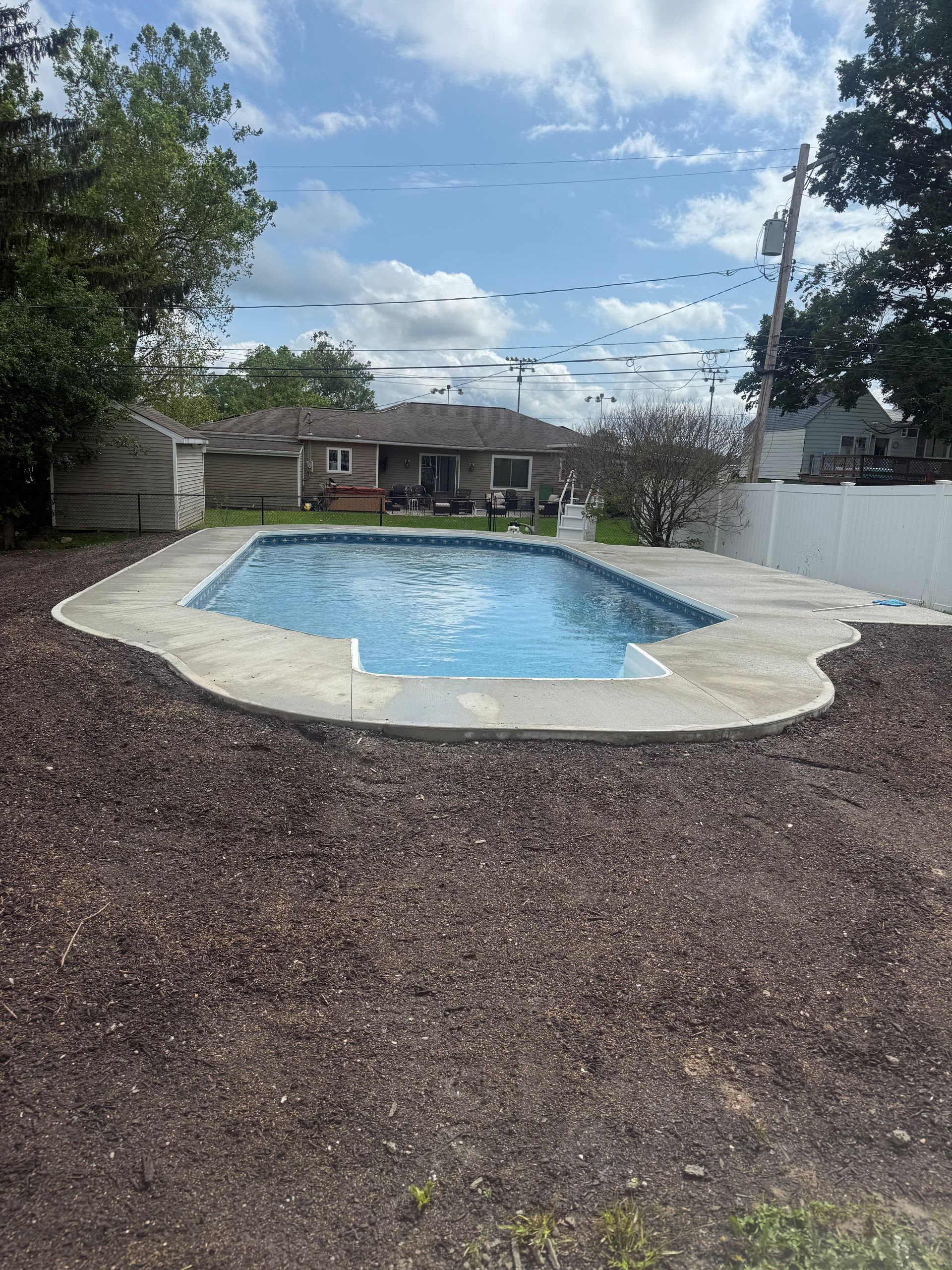 Backyard pool with concrete border, surrounded by mulch, under a cloudy sky.