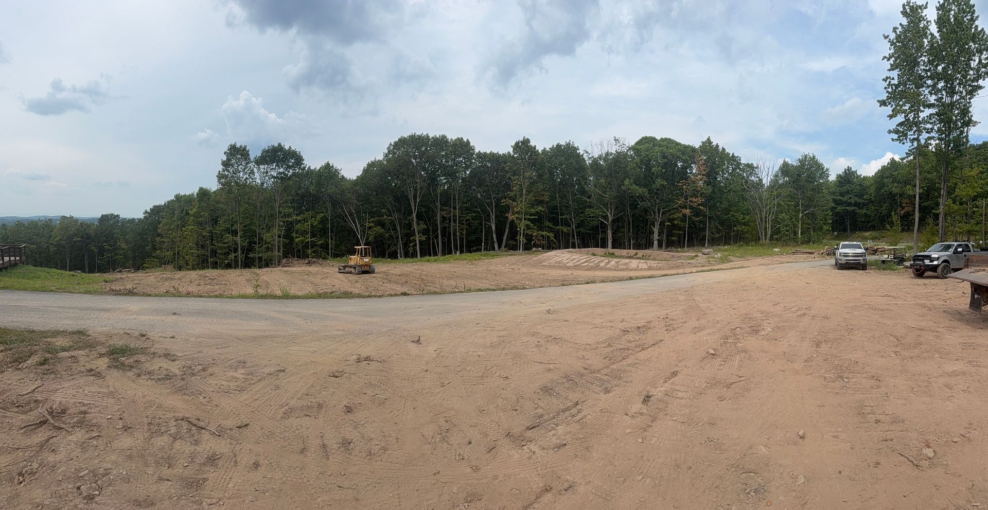 Dirt lot with cleared area, trees in background, cloudy sky.
