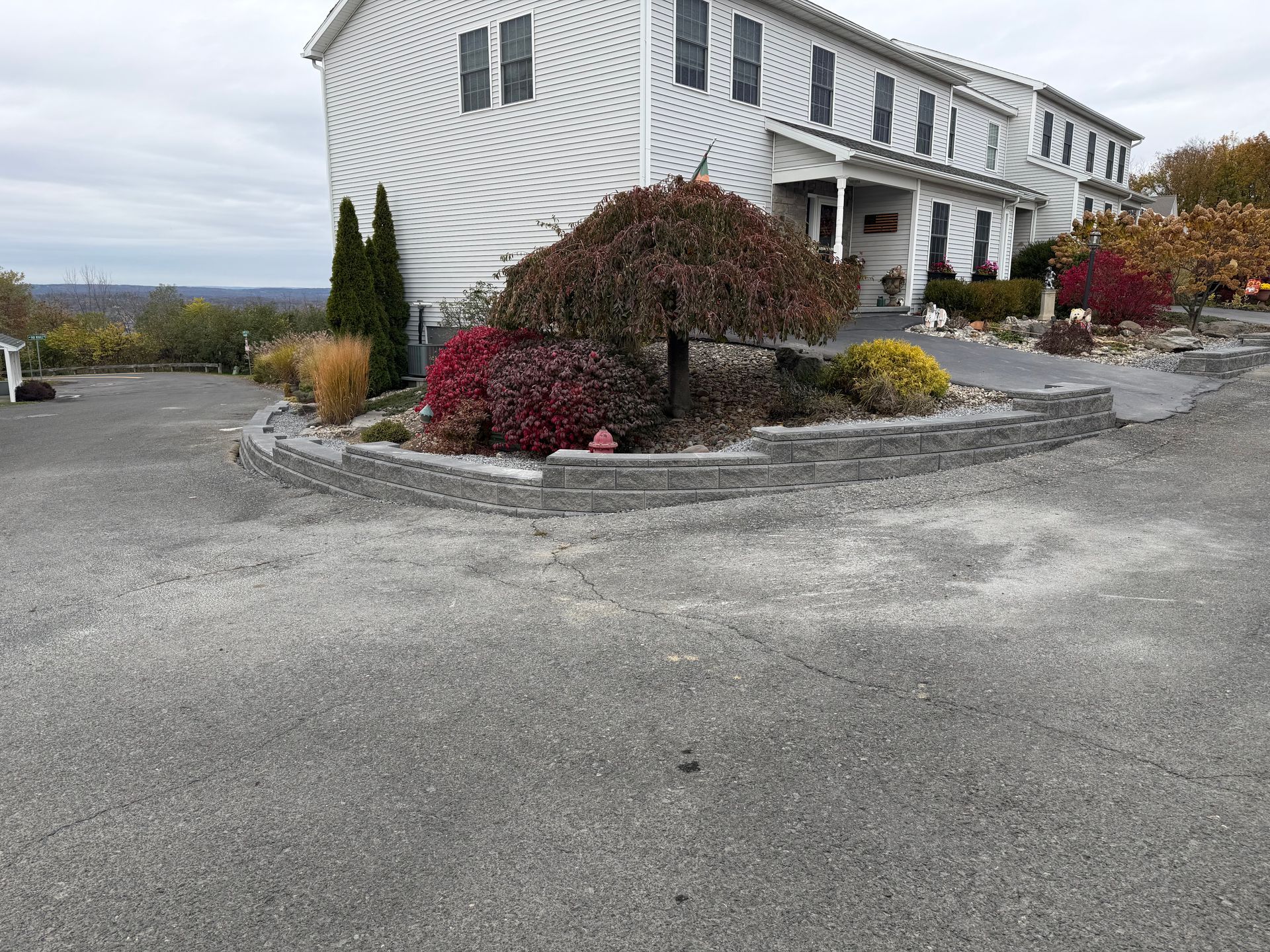 A white house with a stone retaining wall and colorful bushes in front. A driveway leads to the house.