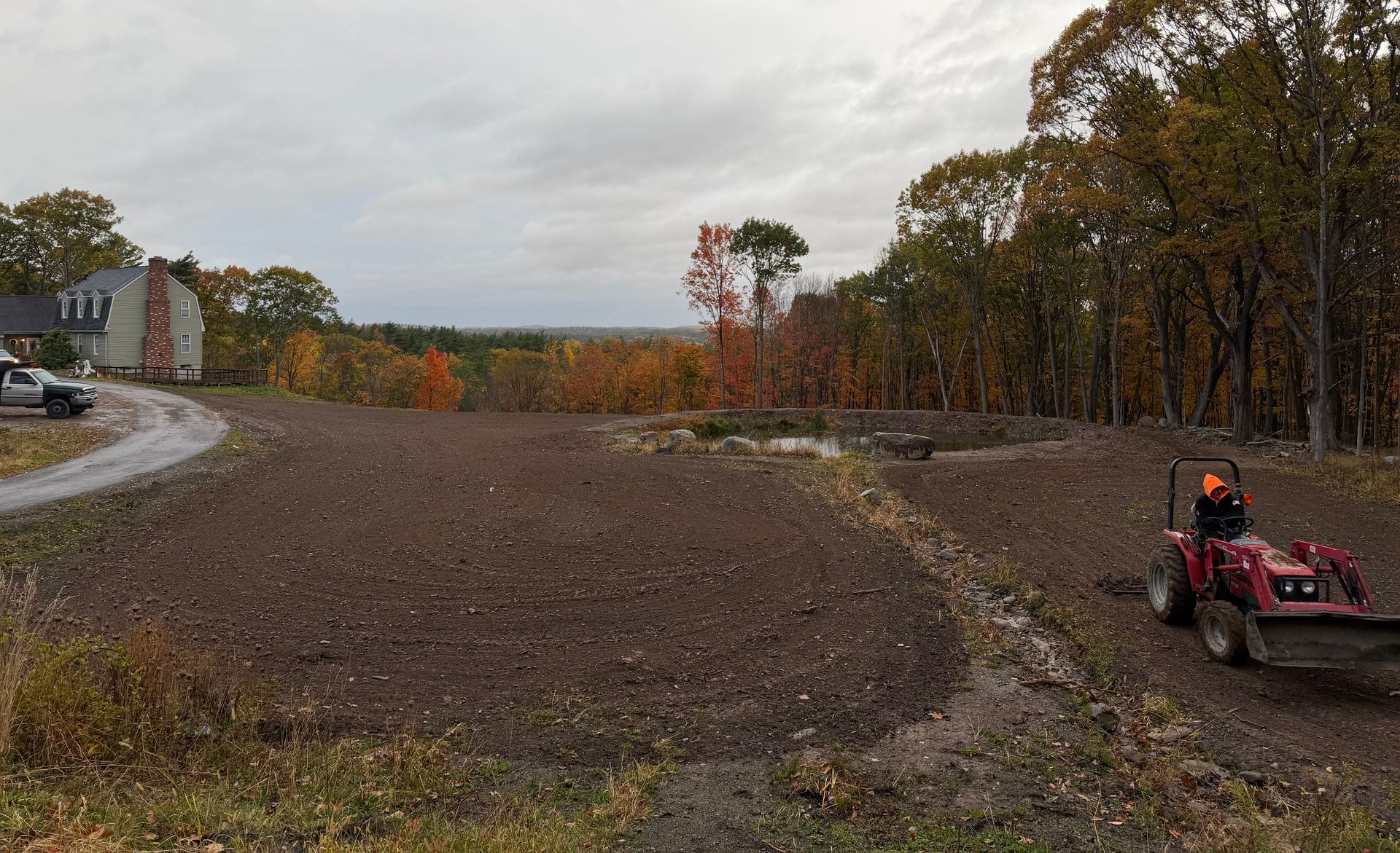 A tractor works on freshly turned earth near a house and trees under an overcast sky.
