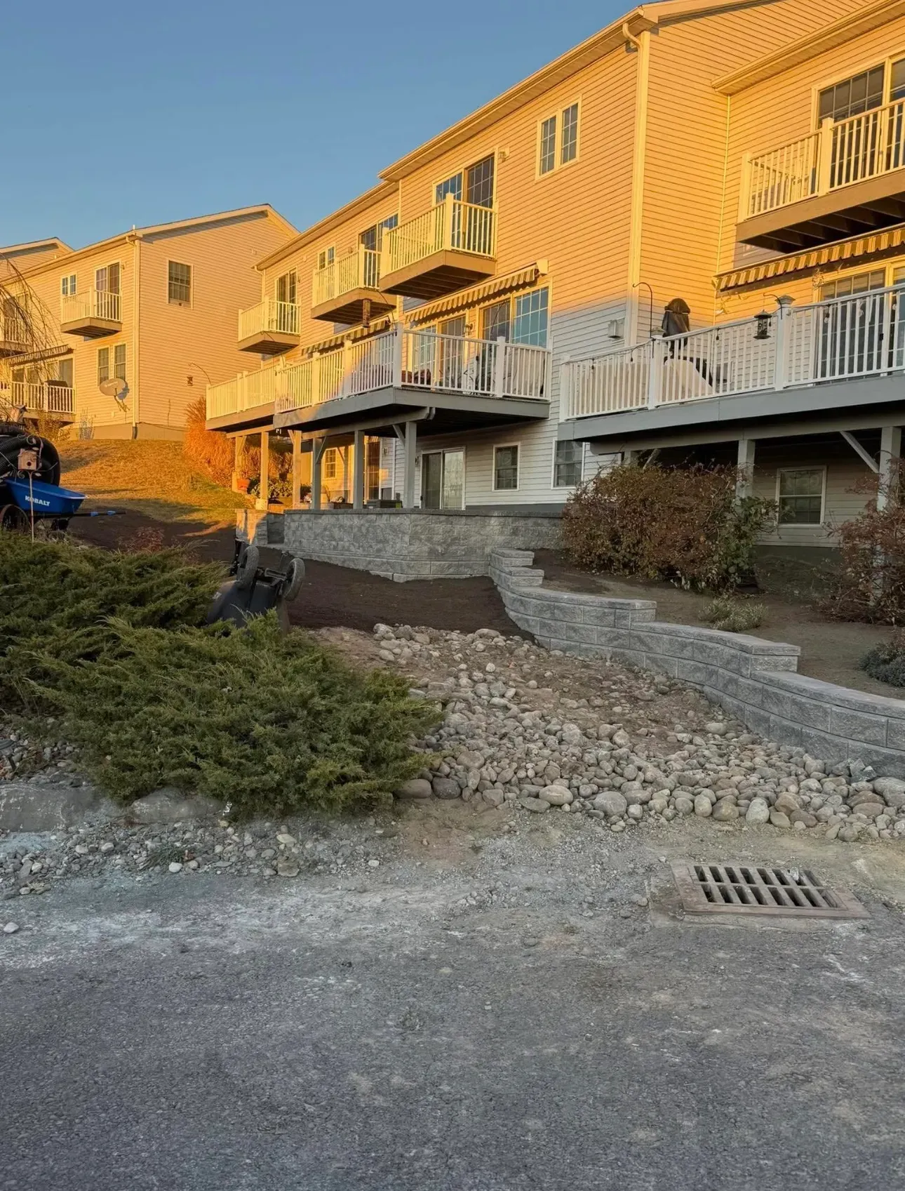 Stone retaining walls tiered on a hillside below multi-story apartment buildings, gravel ground in foreground.