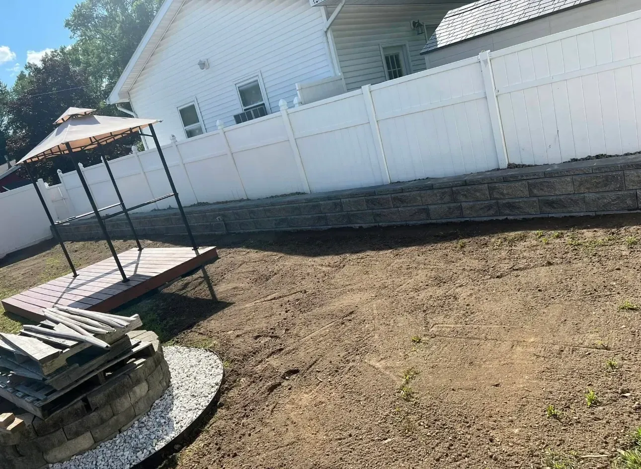 Backyard with dirt ground, gazebo, white fence, and stone retaining wall.
