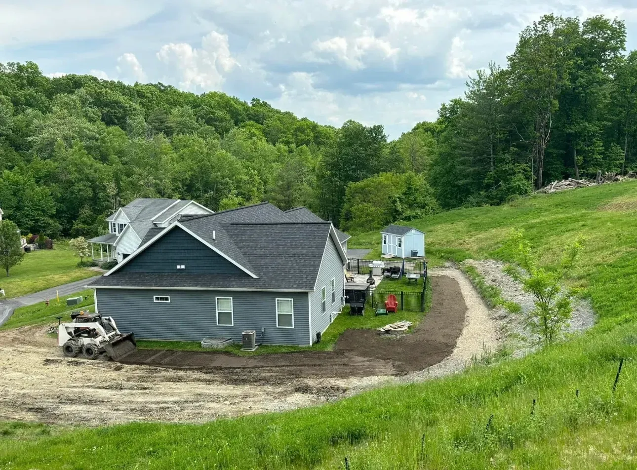 Blue house with construction work in progress on a hillside, gravel path, green grass, and trees in background.