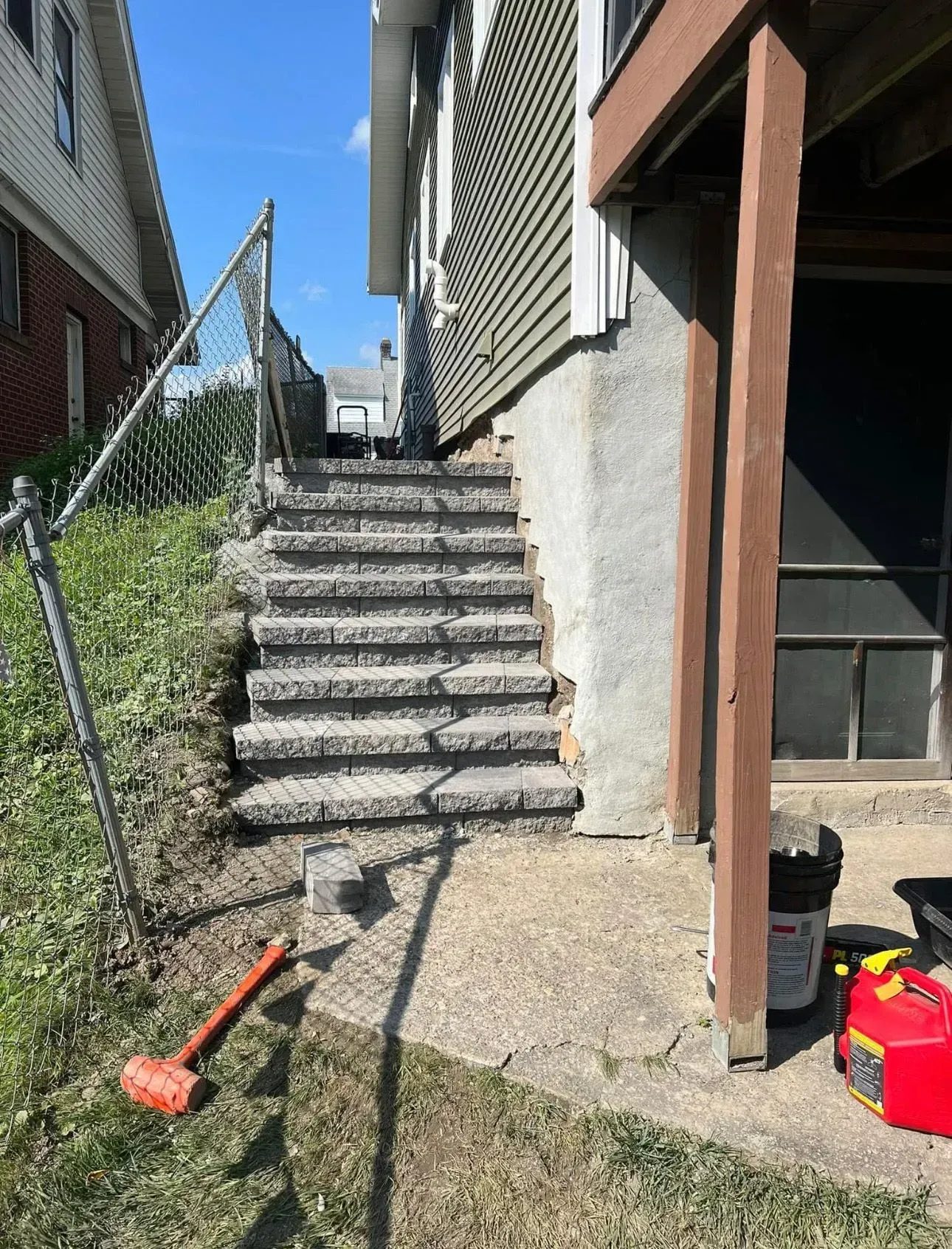 Concrete steps leading up alongside a house with green siding, a fence, and a red sledgehammer on the ground.