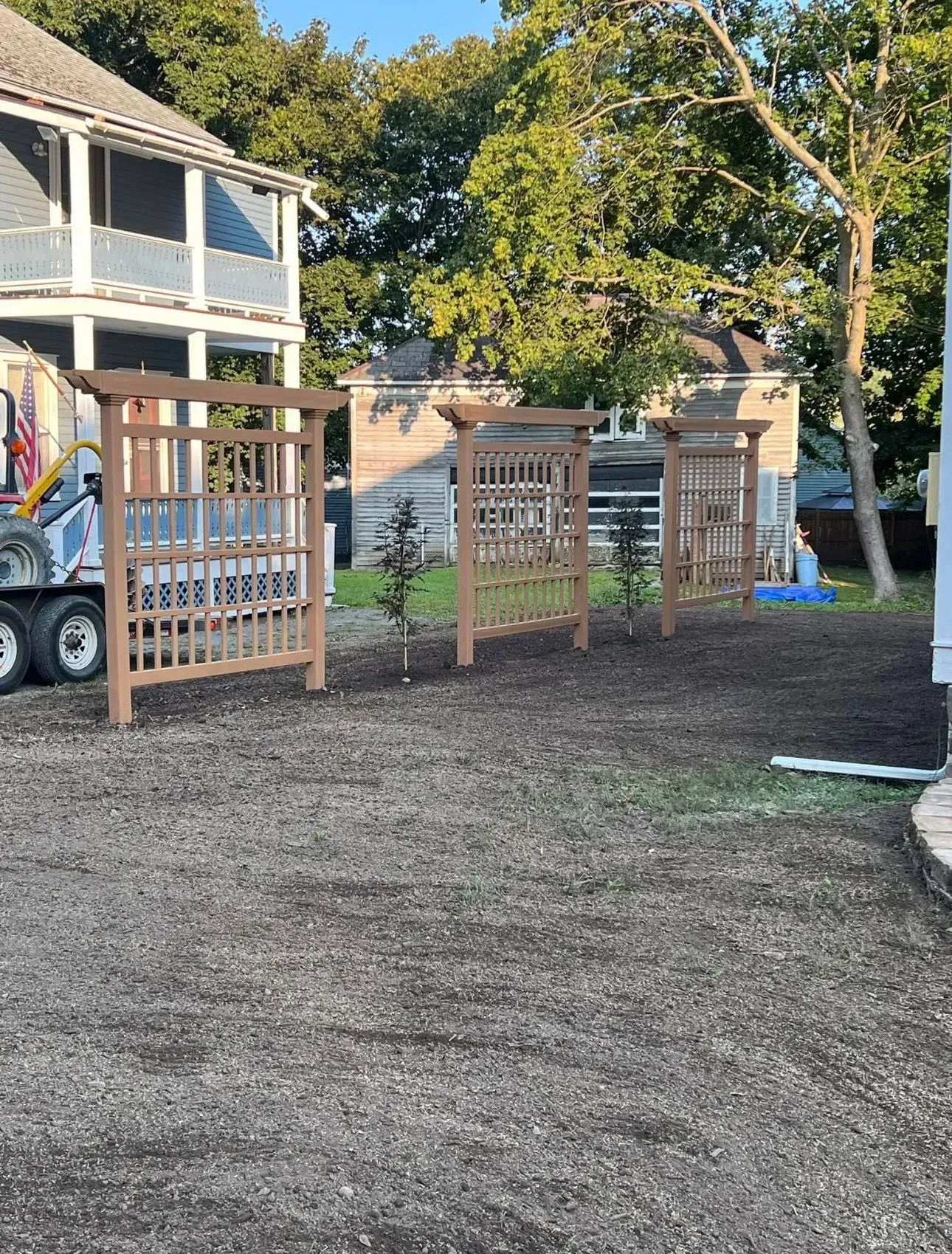 Three brown lattice garden structures set on gravel, small trees between them, near houses and trees.