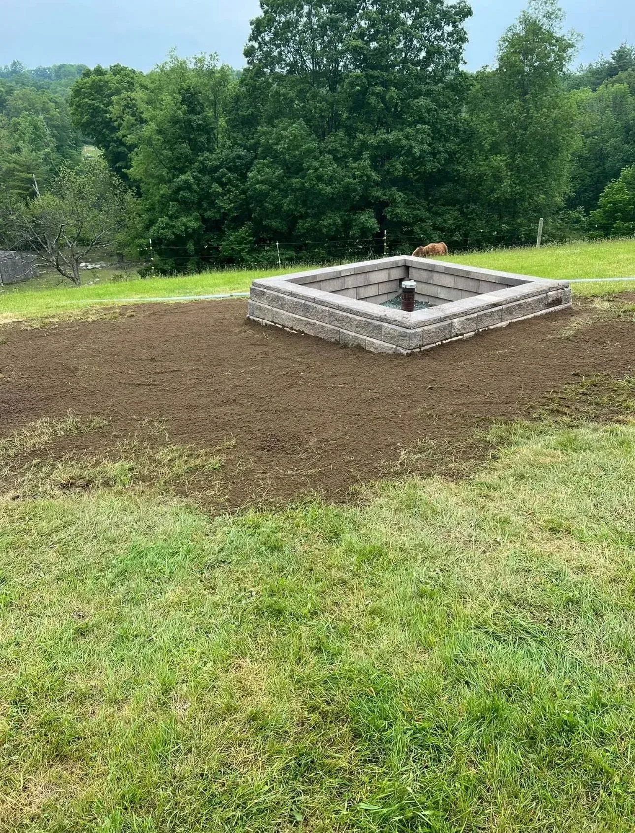 A square brick fire pit on a grassy area, surrounded by dirt, with trees in the background.