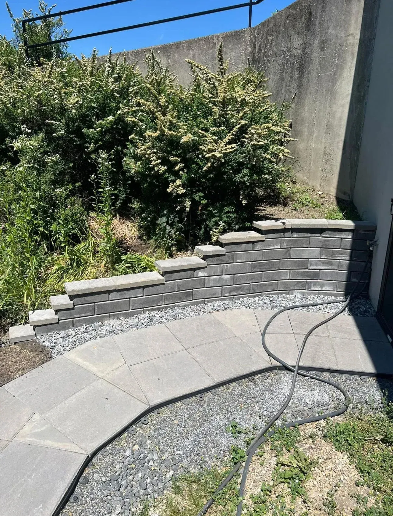 A gray block retaining wall with a stone path, gravel, and bushes under a concrete wall on a sunny day.