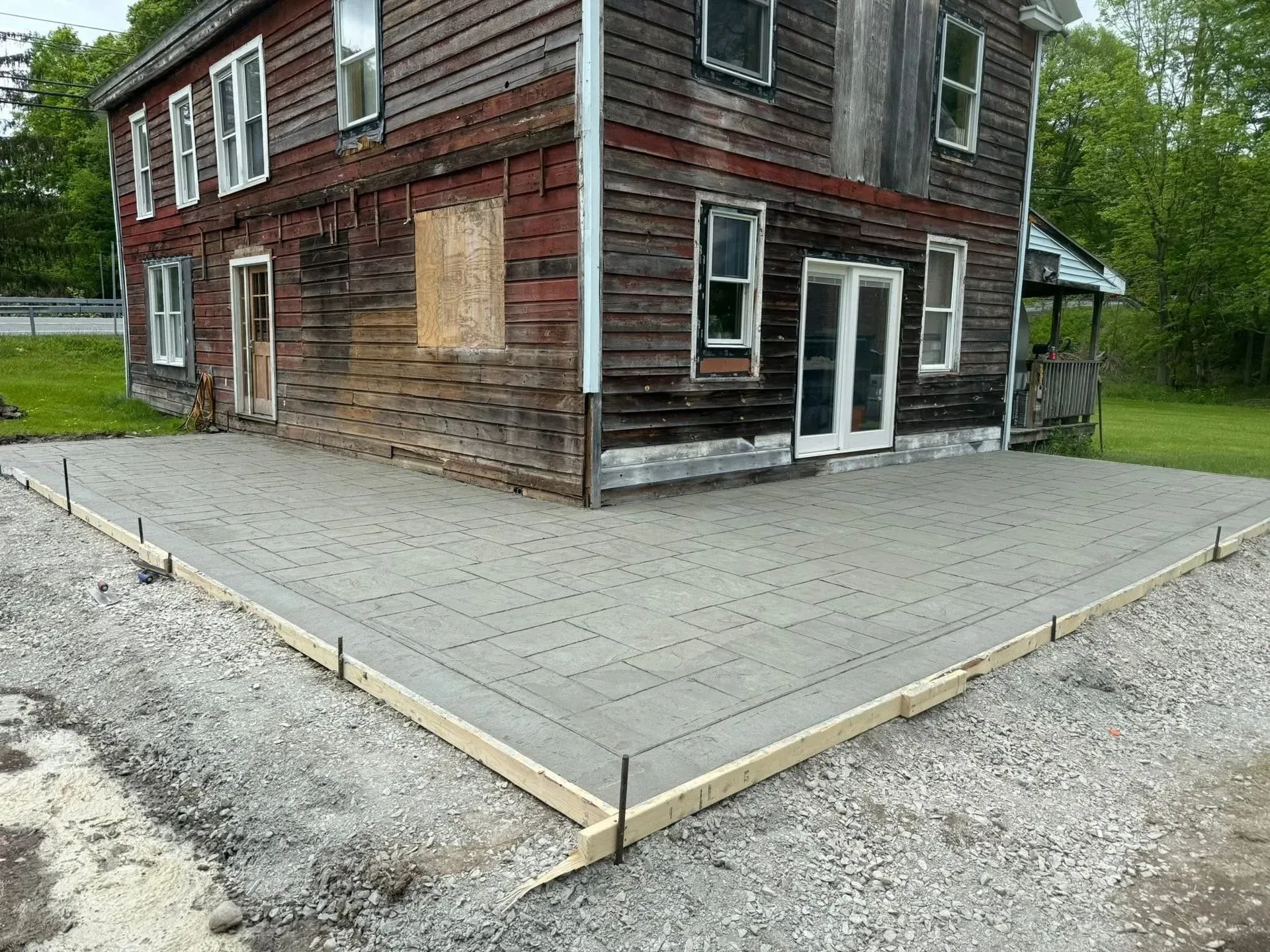 Newly poured concrete patio next to a weathered, wooden house. Gravel borders the patio.