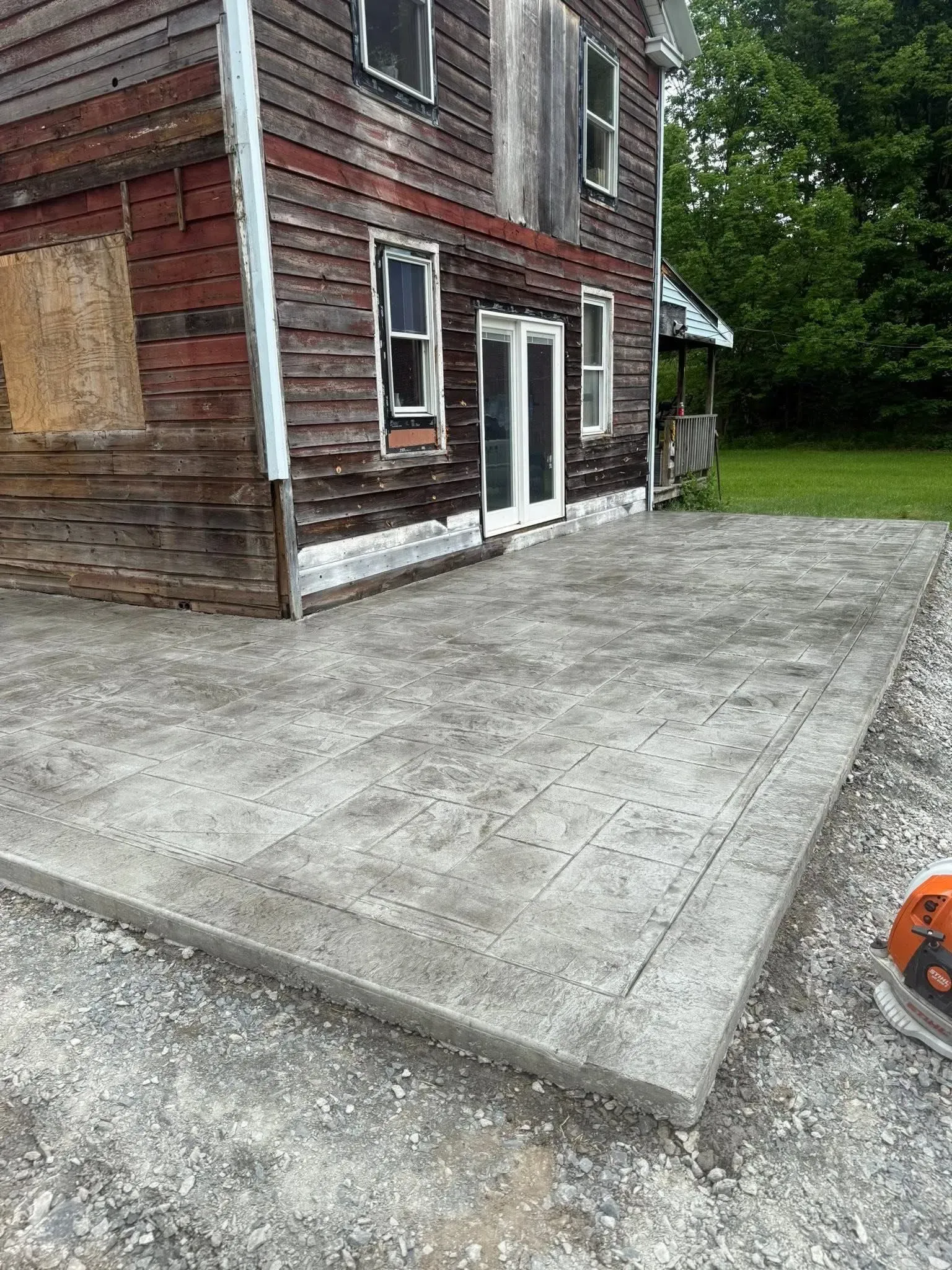 Concrete patio next to a weathered, wooden house with French doors. Gravel border visible.