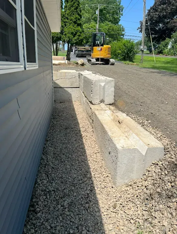 Concrete blocks forming a low wall along a building's side, set on gravel. Mini excavator in the background.