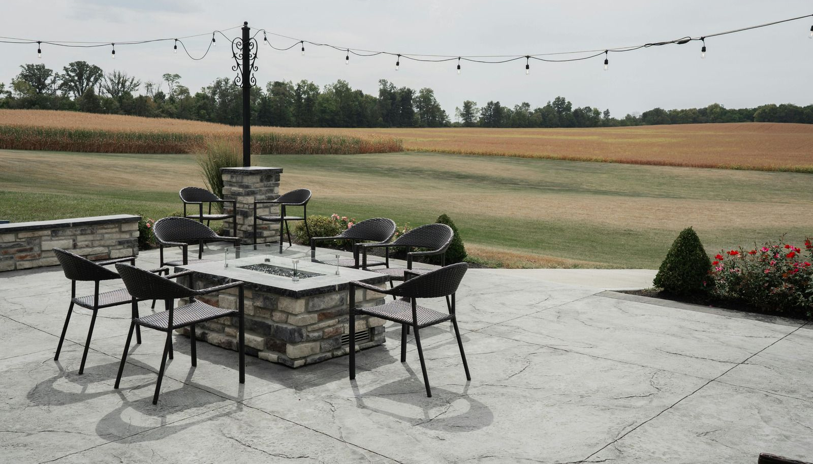 Patio with fire pit and chairs, overlooking a field with trees under a cloudy sky.