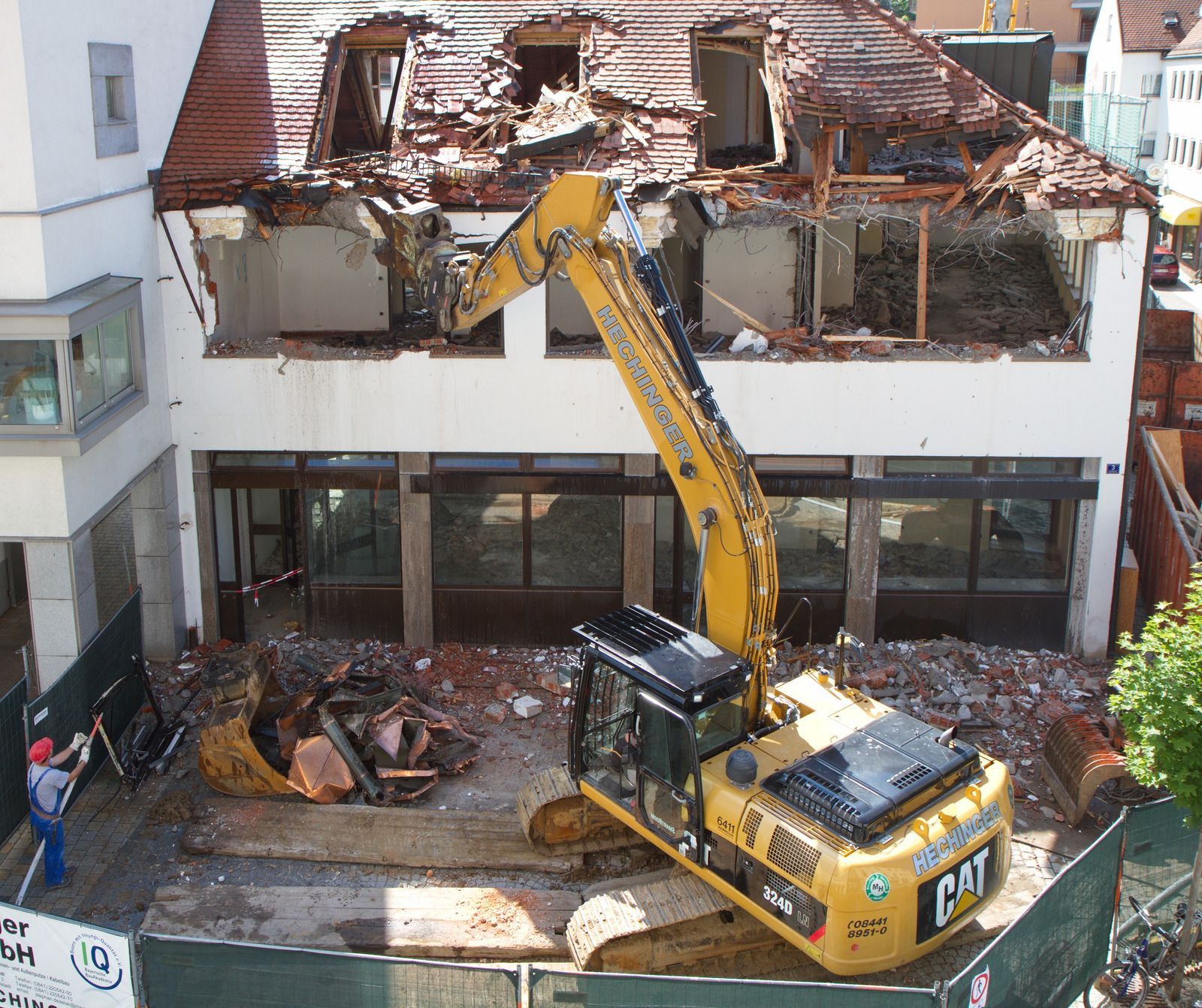 Excavator demolishing a building with a damaged roof and empty windows. A worker stands nearby.