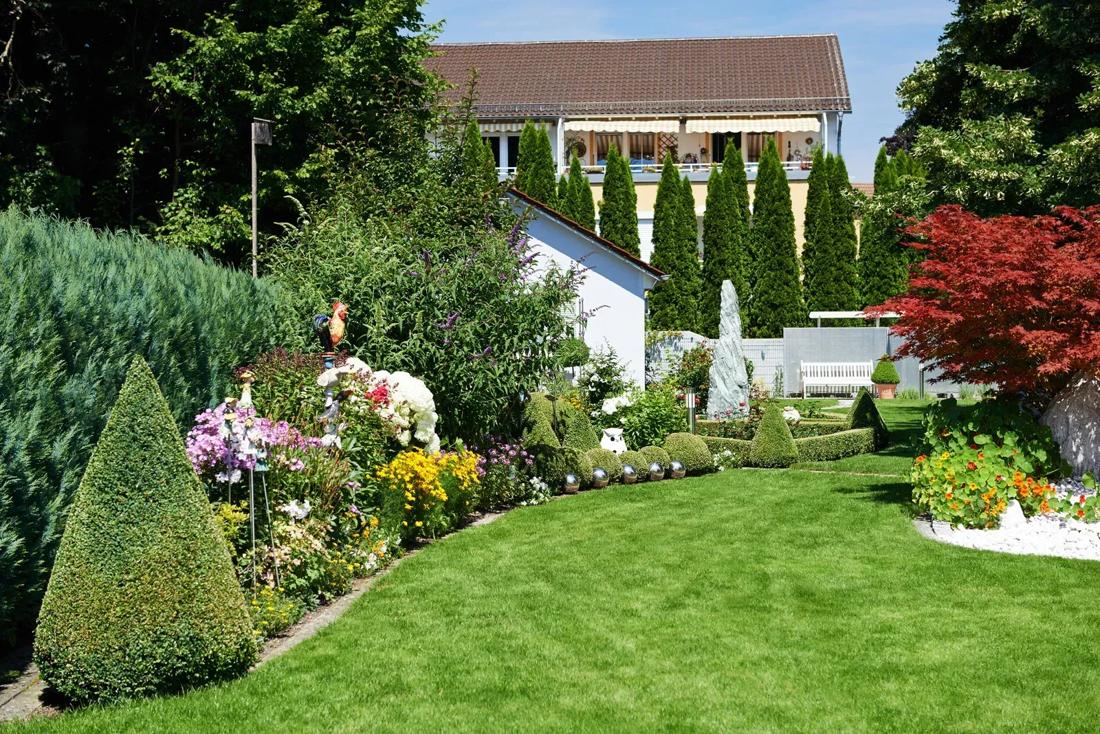 Lush green garden with manicured lawn, flowering plants, and a house in the background.