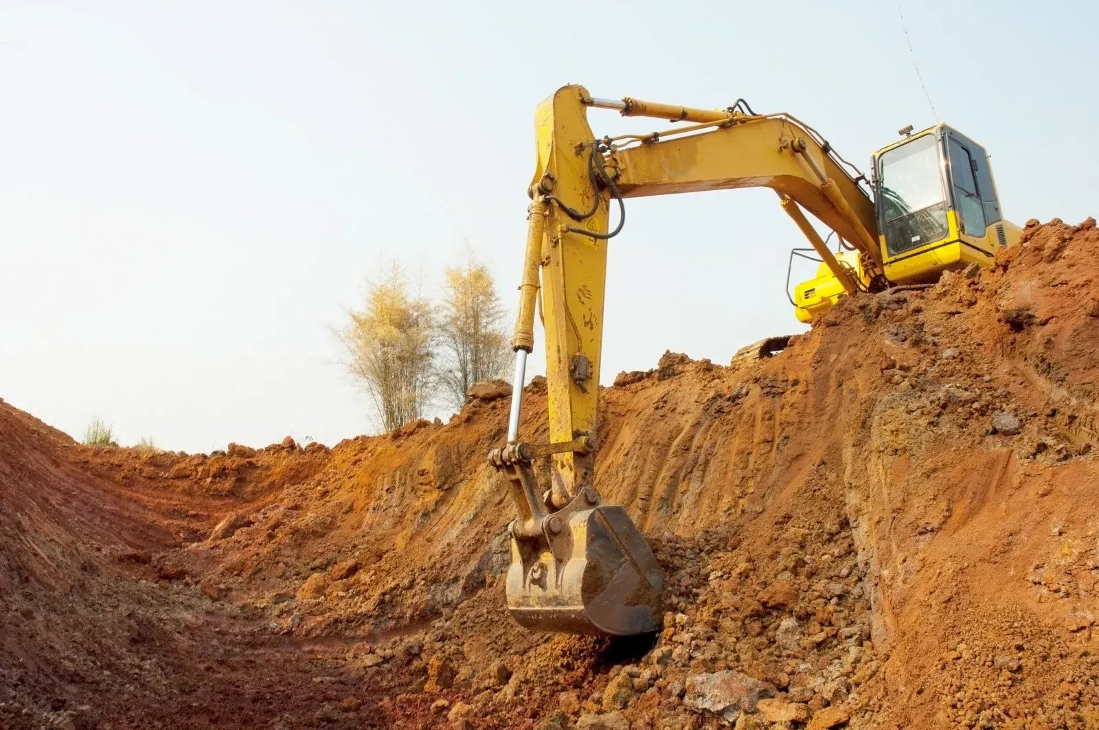 Yellow excavator digging into a reddish-brown earth pile, under a clear sky.