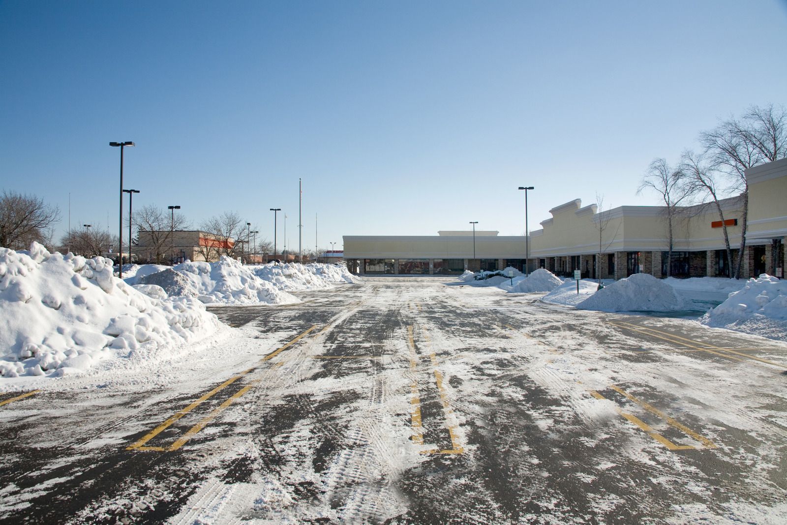 Snow-covered parking lot in front of a strip mall on a clear, sunny day. Snow banks line the parking spaces.