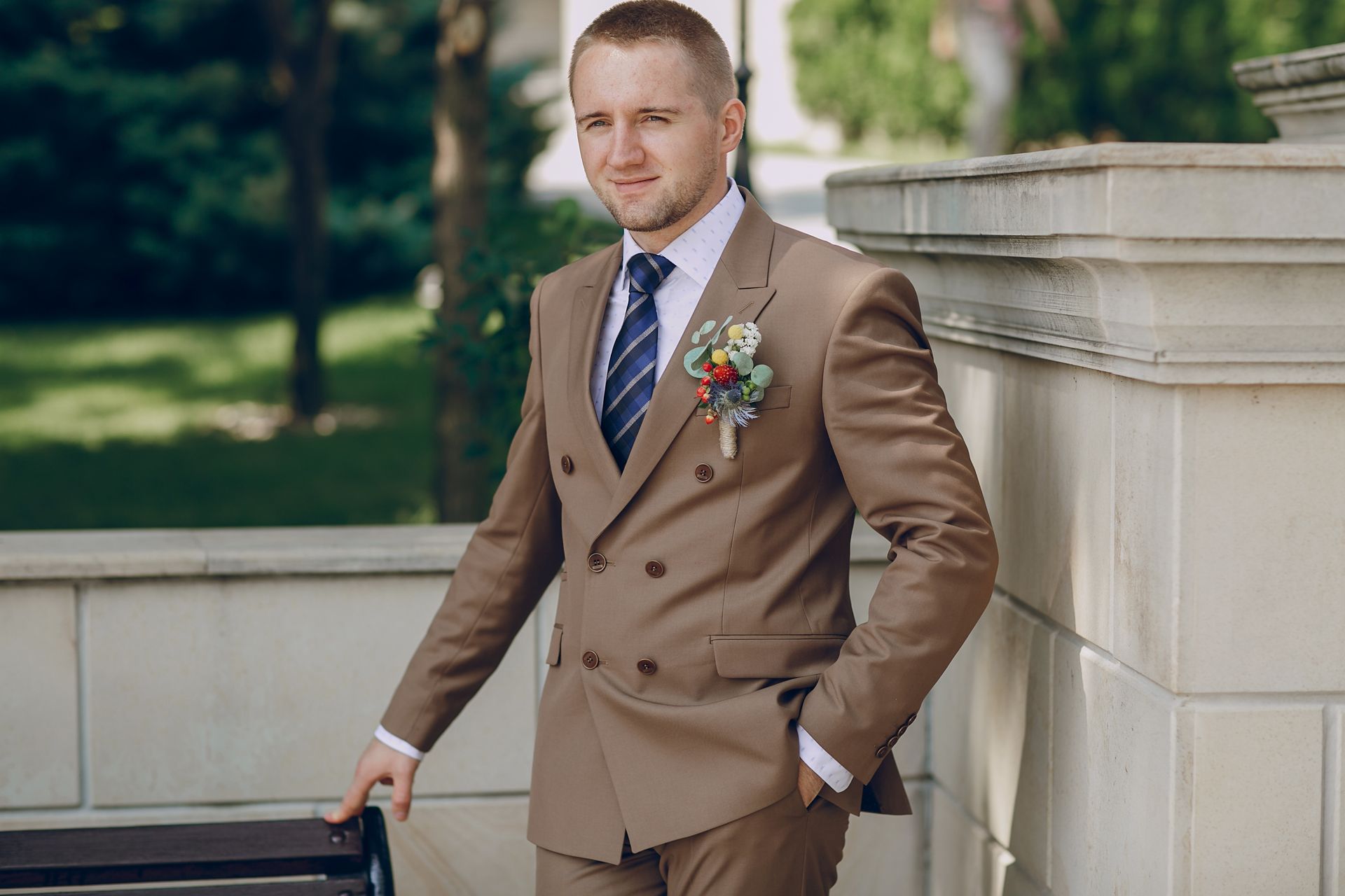 Man in brown suit with tie and boutonniere leans against a wall outside.
