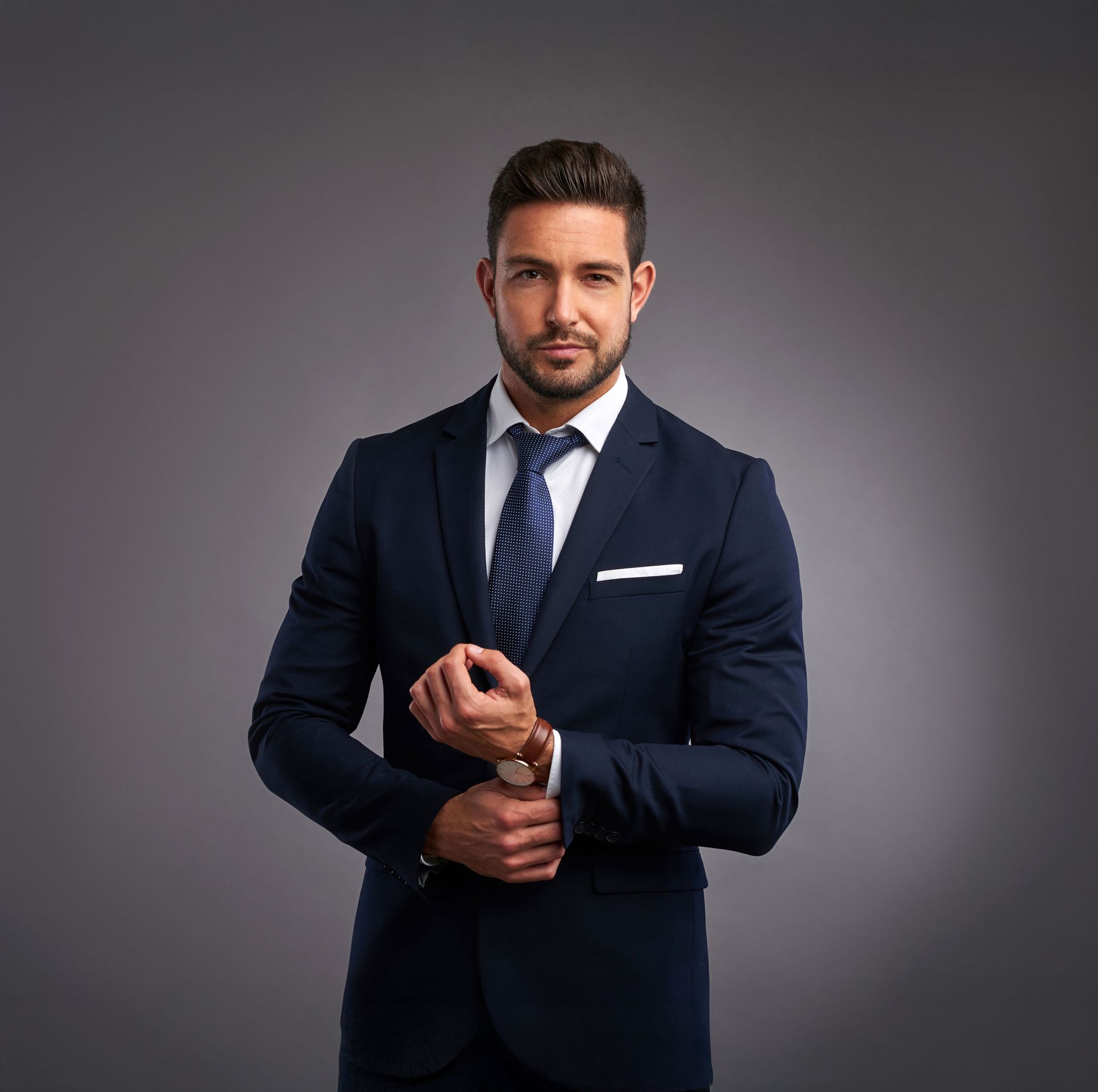 Man in navy suit adjusting cuff, looking at camera against a gray background.