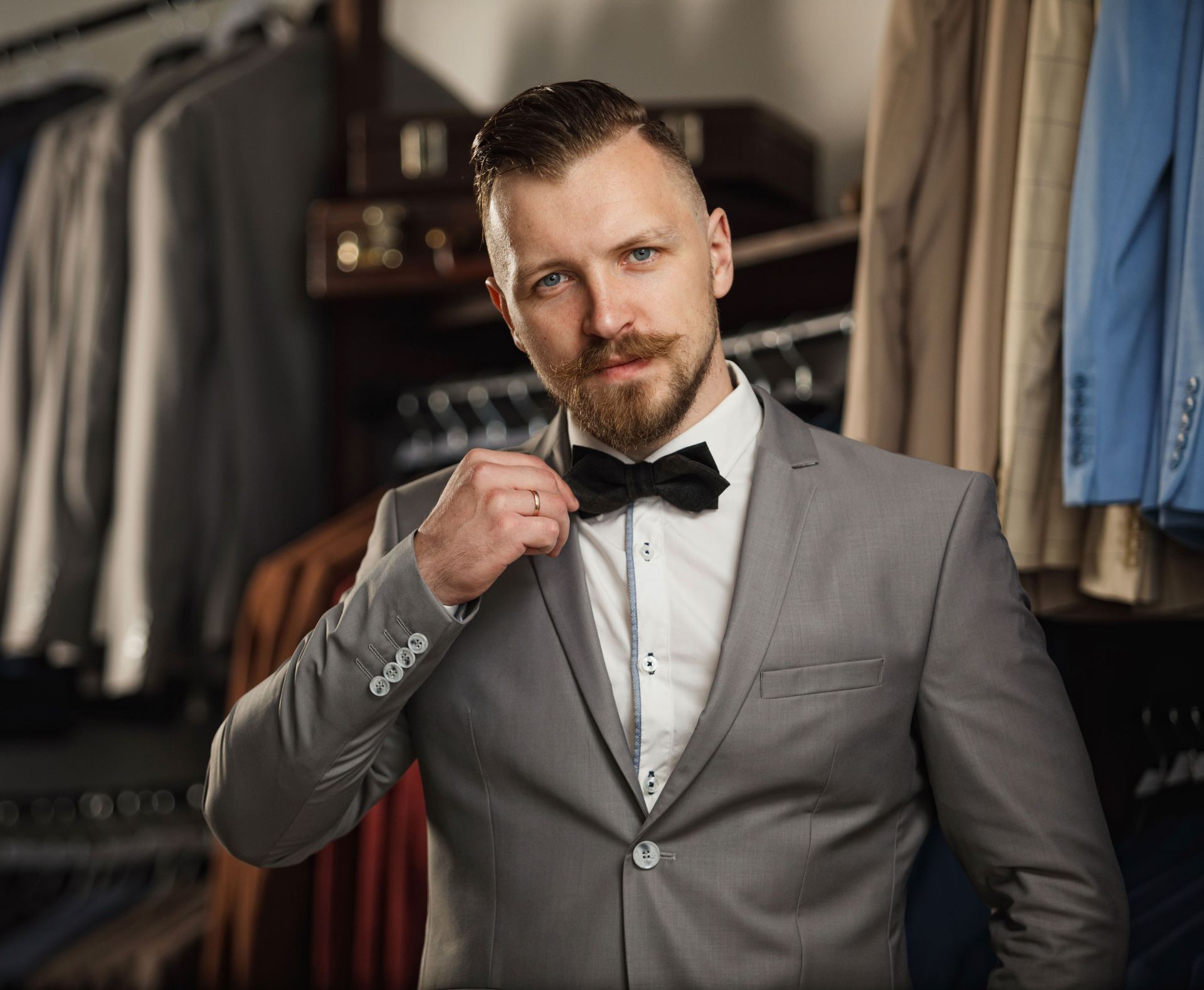 Man in gray suit adjusts bow tie in a suit shop.