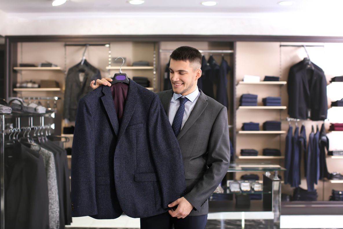 Man in suit holds a navy blue patterned blazer in a clothing store, smiling.
