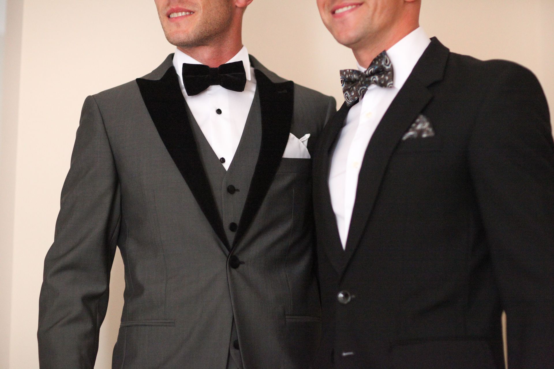 Two men in formal suits with bow ties and pocket squares. One is wearing a gray suit, the other, black.