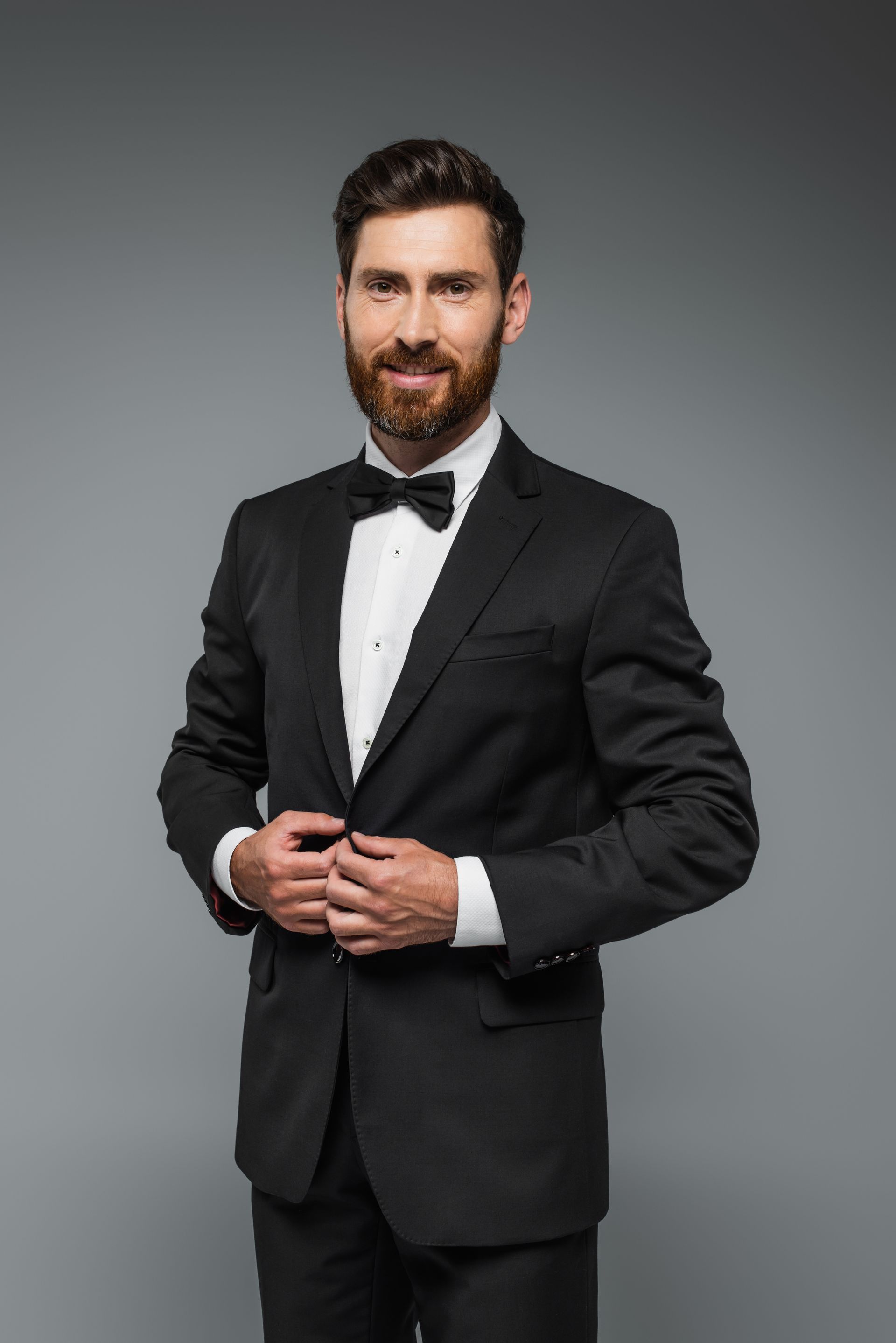 Man in a black tuxedo with a bow tie, smiling, gray background.