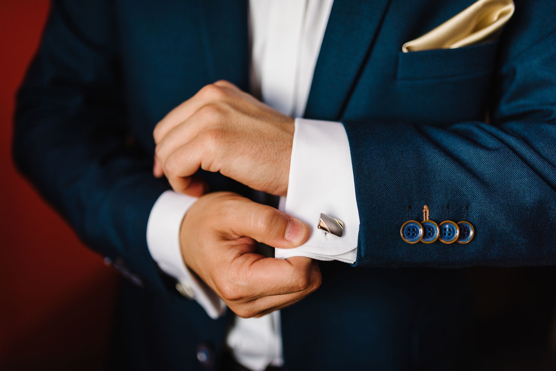 Man adjusting cufflink on a blue suit jacket. White shirt and gold pocket square visible.