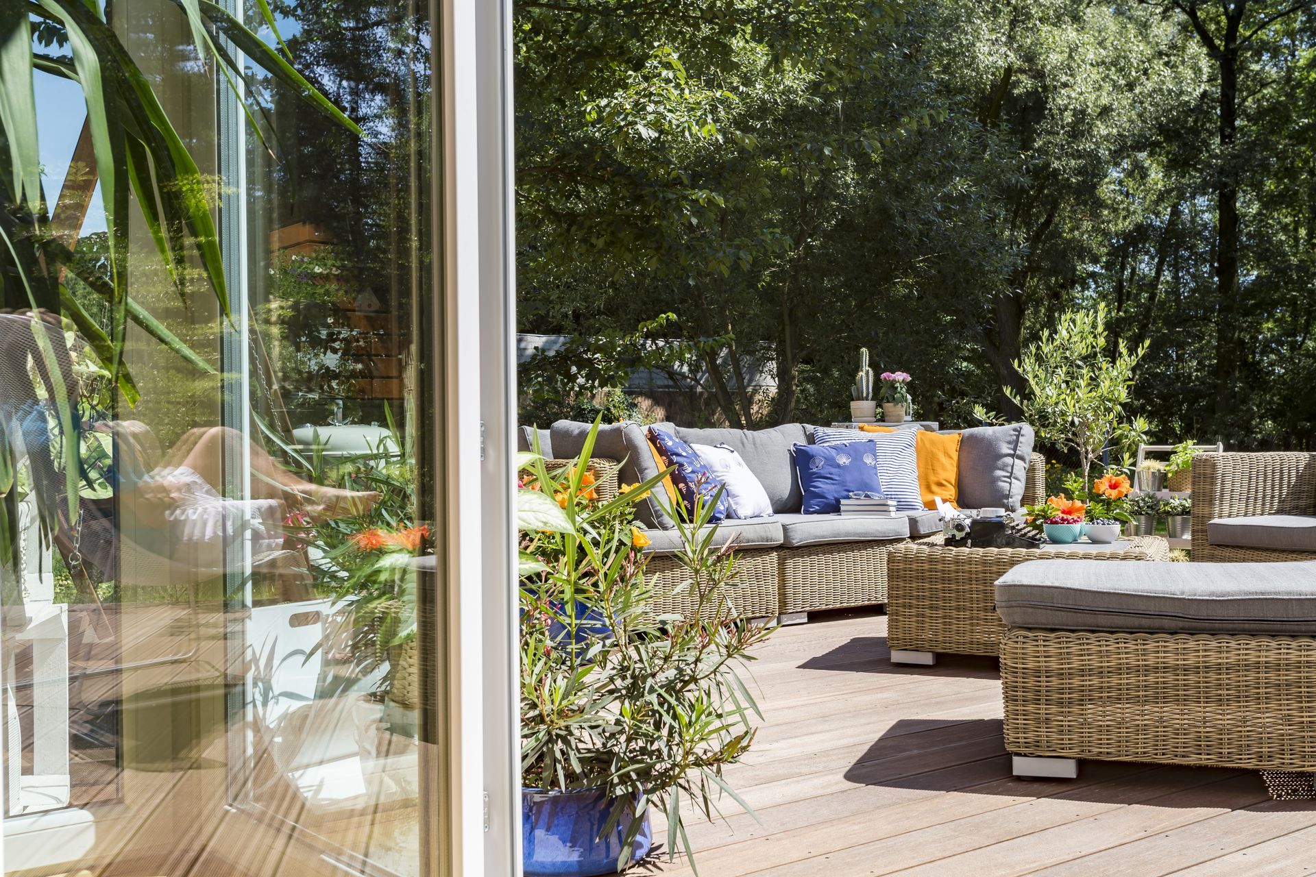 Patio with wicker furniture, cushions, and potted plants overlooking trees, visible through a glass door.