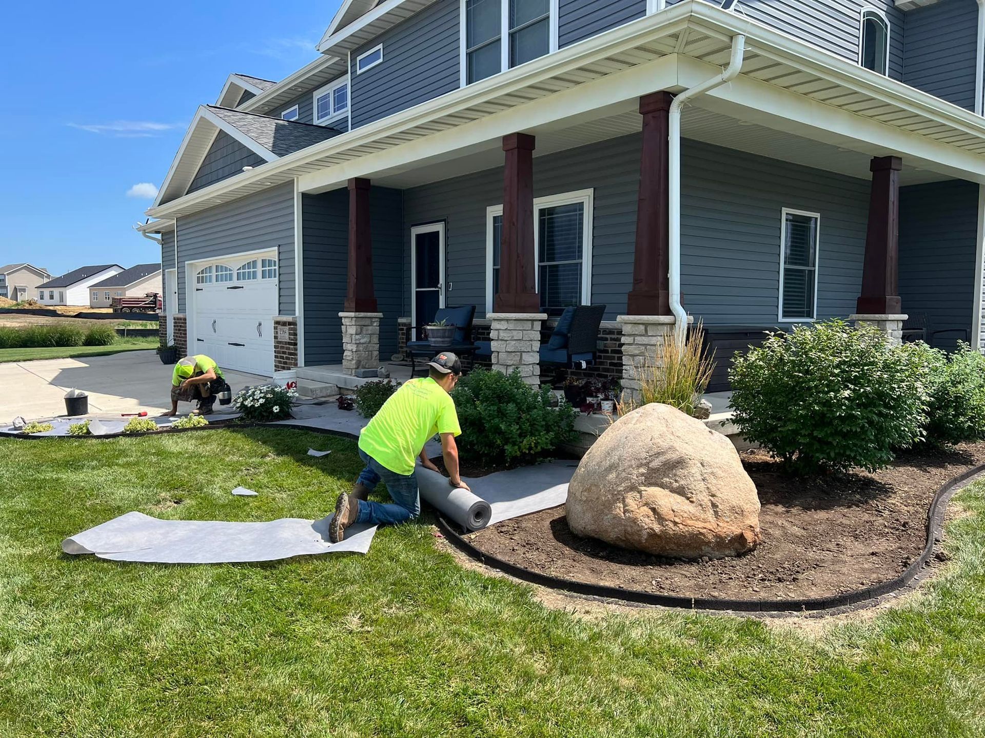 Two workers installing landscaping fabric near a house with a porch and a large rock in a garden bed.