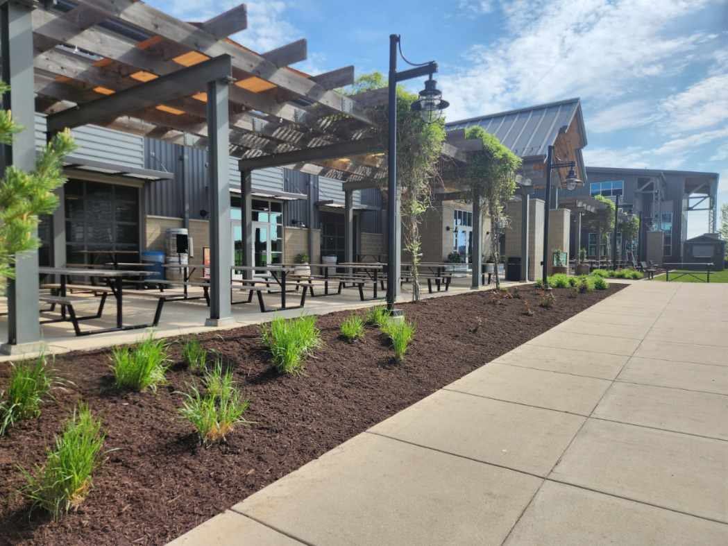 Outdoor dining area with pergola, tables, and landscaping beside a paved walkway, under a blue sky.