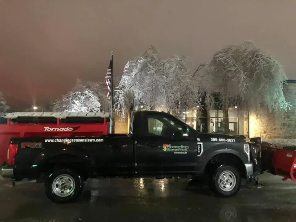 Black pickup truck with snow plow, red salt spreader, icy trees in background.