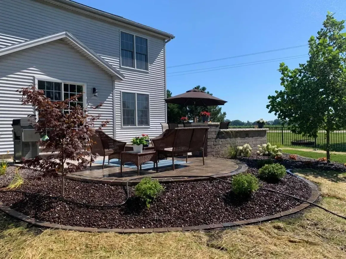 Backyard patio with seating, stone wall, and landscaping, against a two-story house under a blue sky.