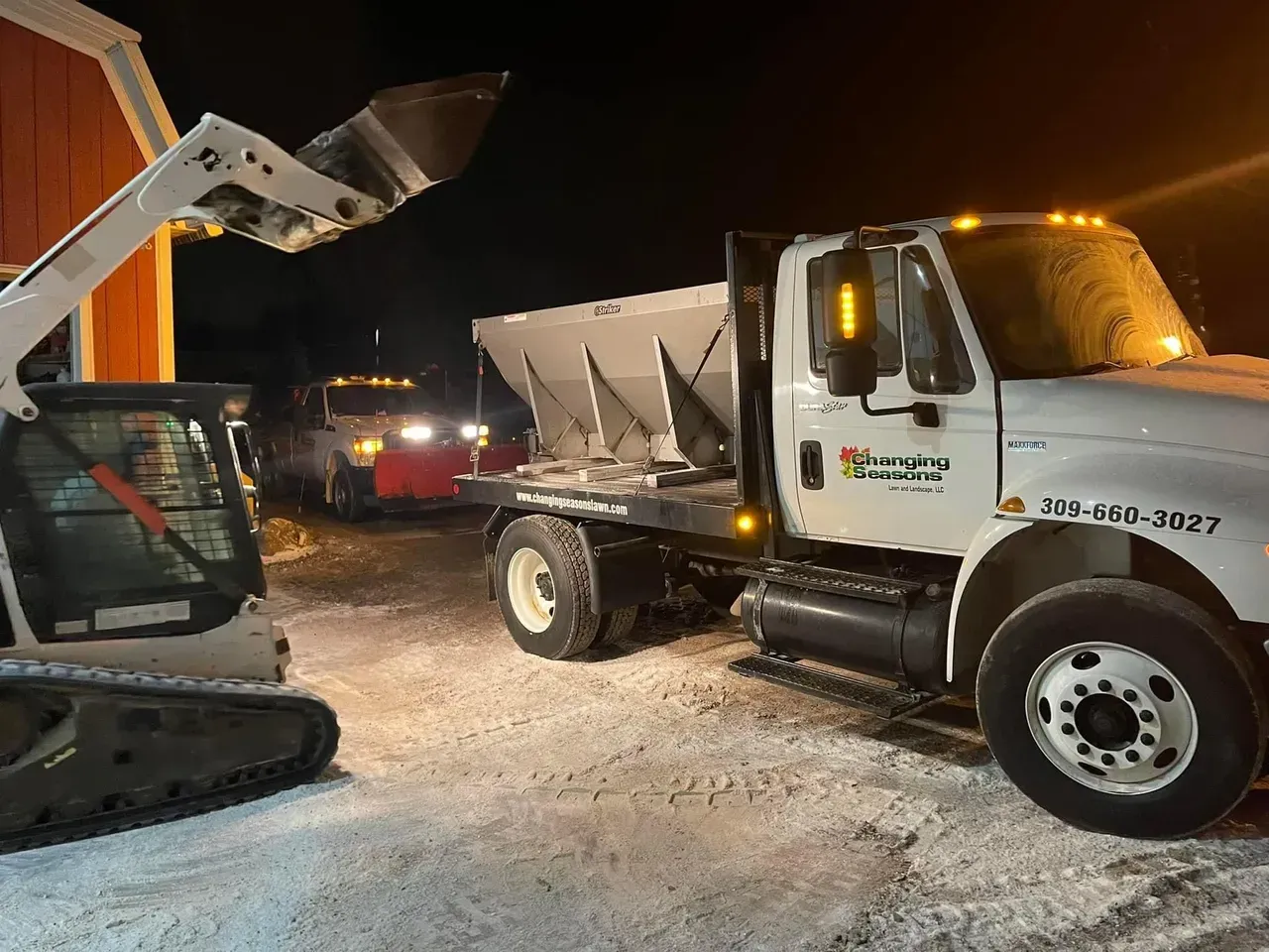 A skid steer loading a snow plow truck with salt at night near a red building.