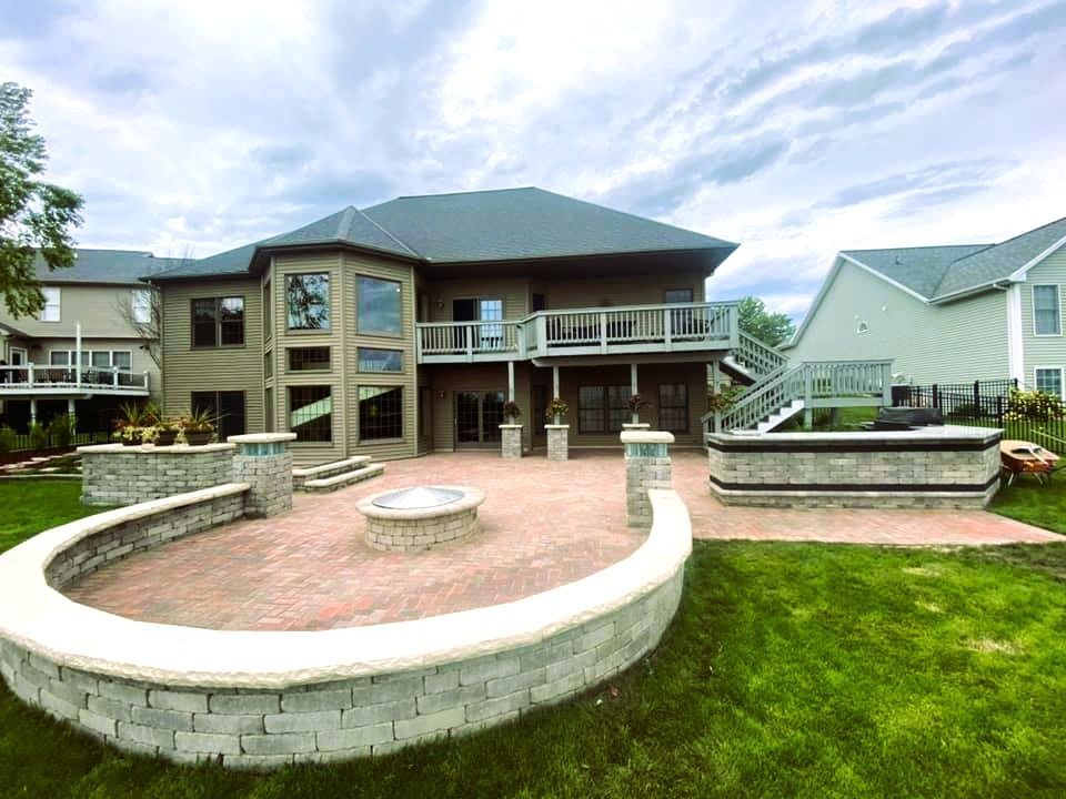 Backyard patio with fire pit, bordering a large house with two levels and a deck. Green grass and overcast sky.