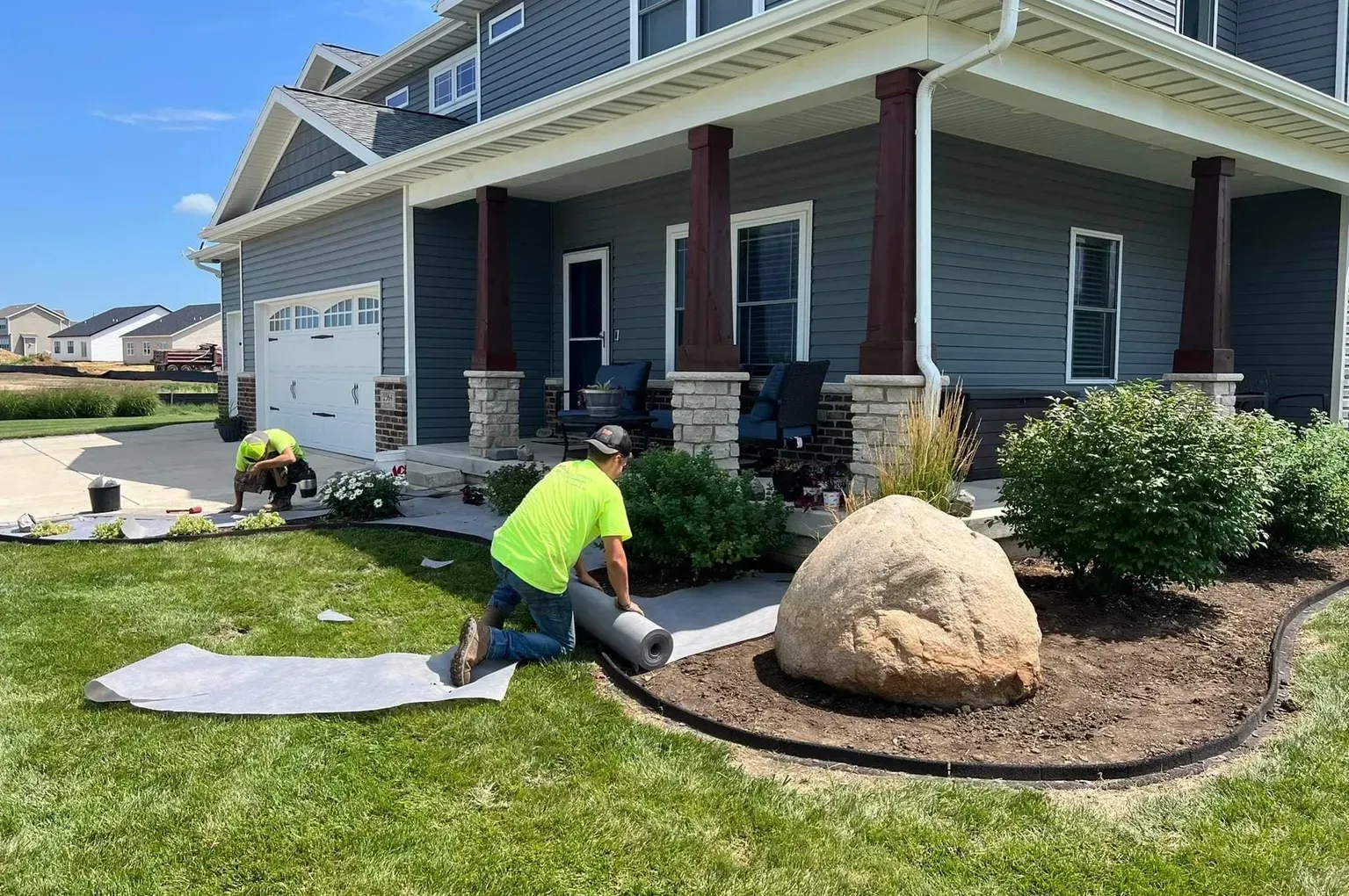 Two workers installing landscaping fabric near a house. One kneels rolling the fabric, other works nearby.
