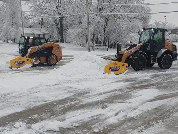 Two orange snowplows clearing snow from a parking lot on a snowy day.