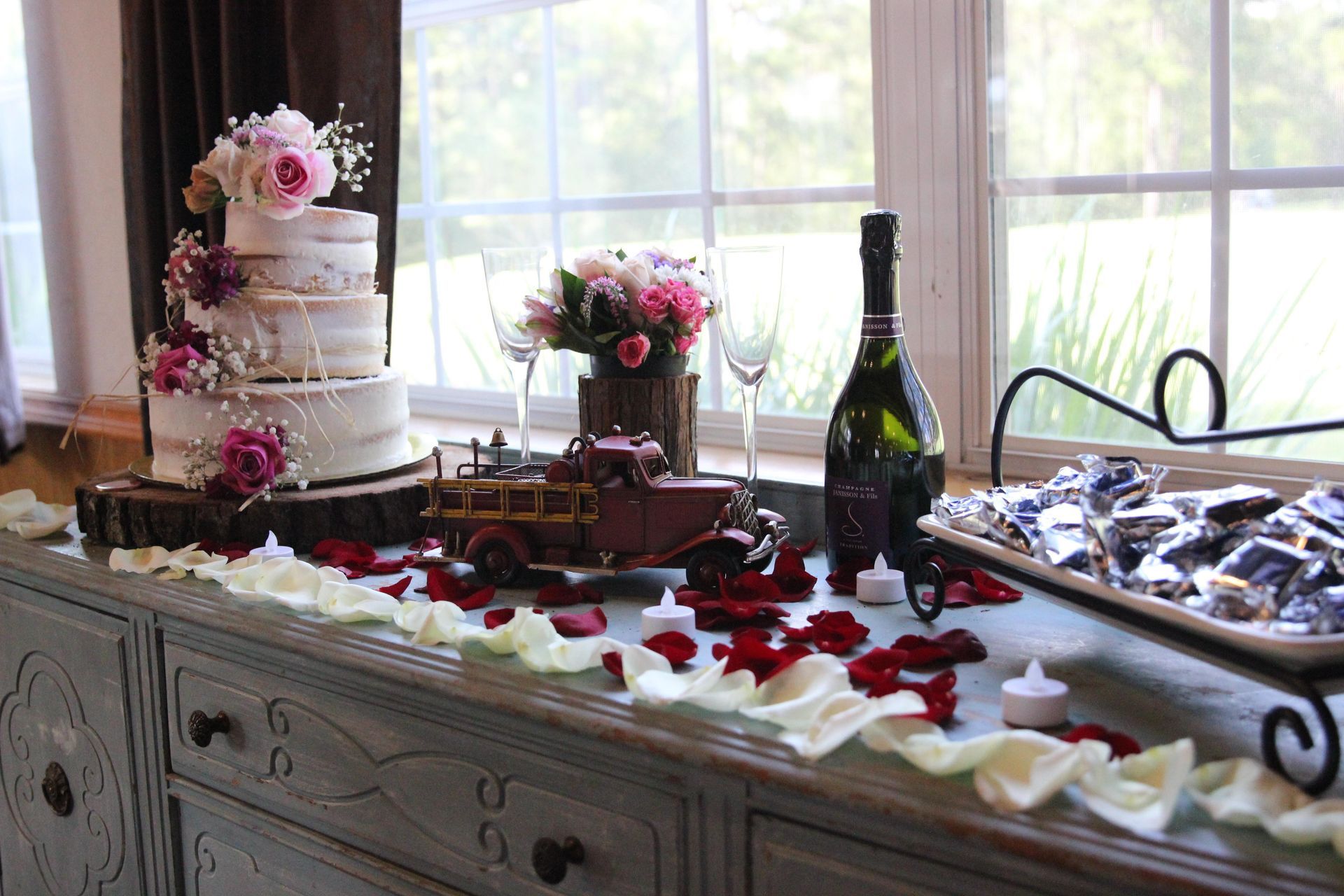 A table with a wedding cake , a bottle of wine , candles , and a toy truck.