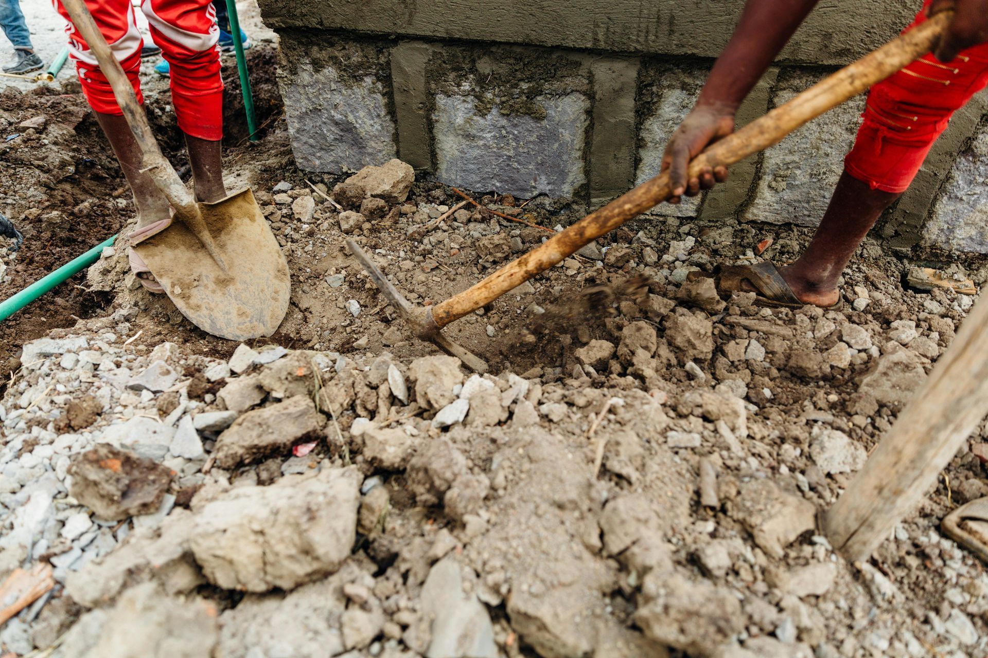 Two people in red pants dig soil with a shovel and hoe near a stone structure.