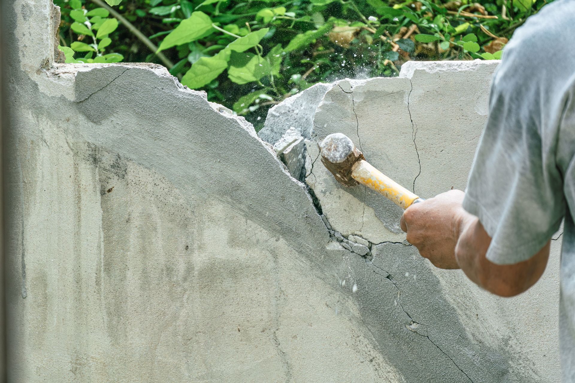 Person in a gray shirt using a sledgehammer to break a gray concrete wall outside.