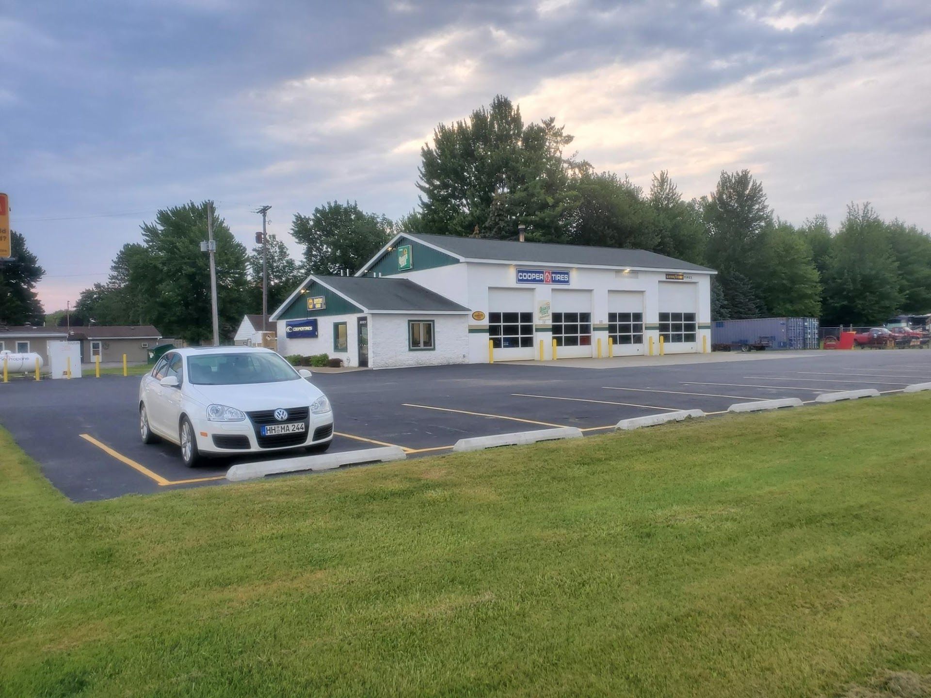 White car parked in front of auto repair shop with green trim; asphalt parking lot, green grass, and trees.