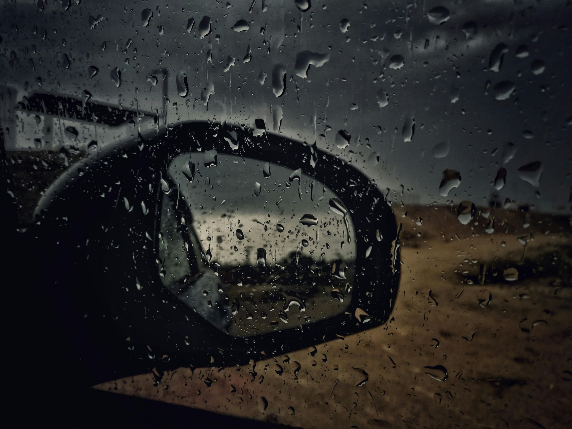 Raindrops on a car window; side mirror reflecting a dark, overcast landscape.