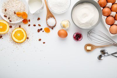 Baking ingredients arranged on a white surface: flour, eggs, fruit, butter, and kitchen tools.