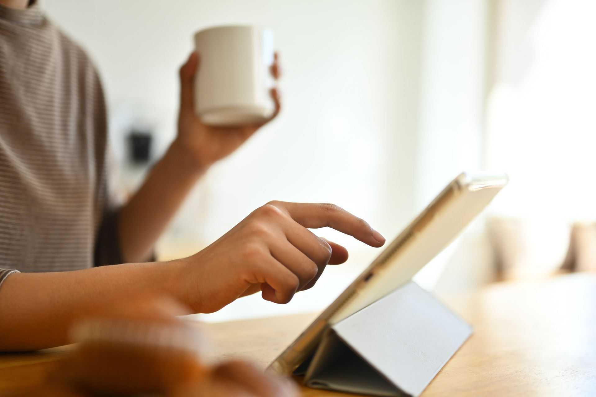 Person using a tablet with one hand and holding a mug with the other hand at a table.