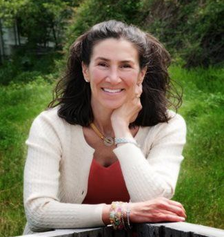 Woman smiles, resting chin on hand, outdoors; wearing white sweater, orange top, jewelry.