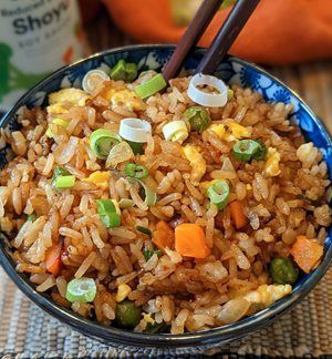 Bowl of fried rice with peas, carrots, and green onions. Chopsticks on top, soy sauce in background.