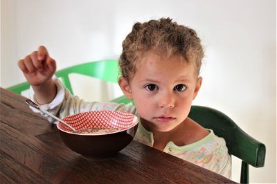 Girl at a table with cereal, raising a hand, looking at the camera.
