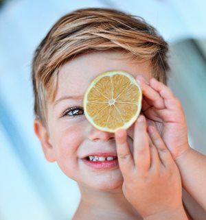 Young child smiling, holding lemon slice over one eye.