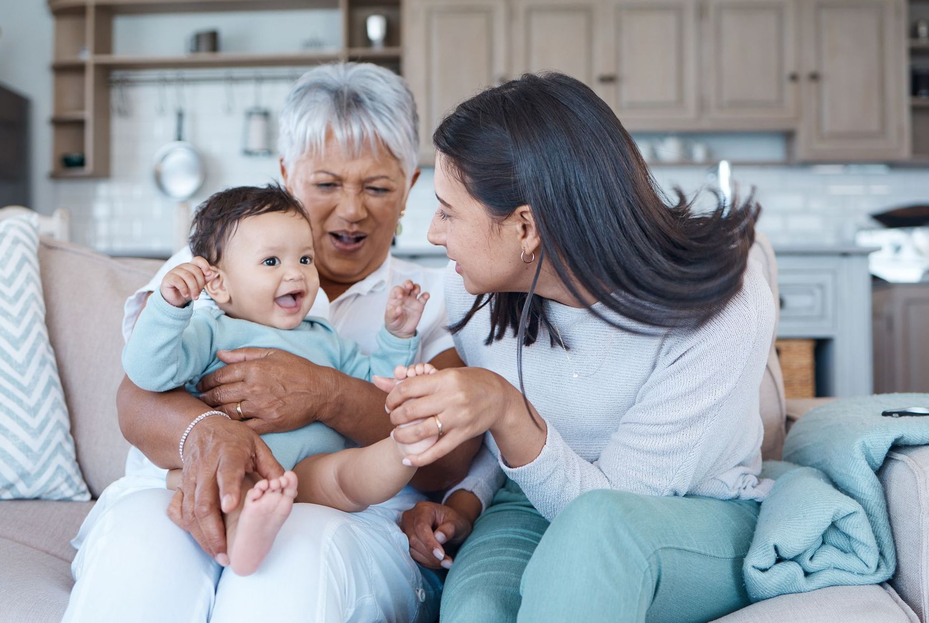Grandmother, mother, and baby on a couch; baby laughs, mother touches feet, and grandmother holds.