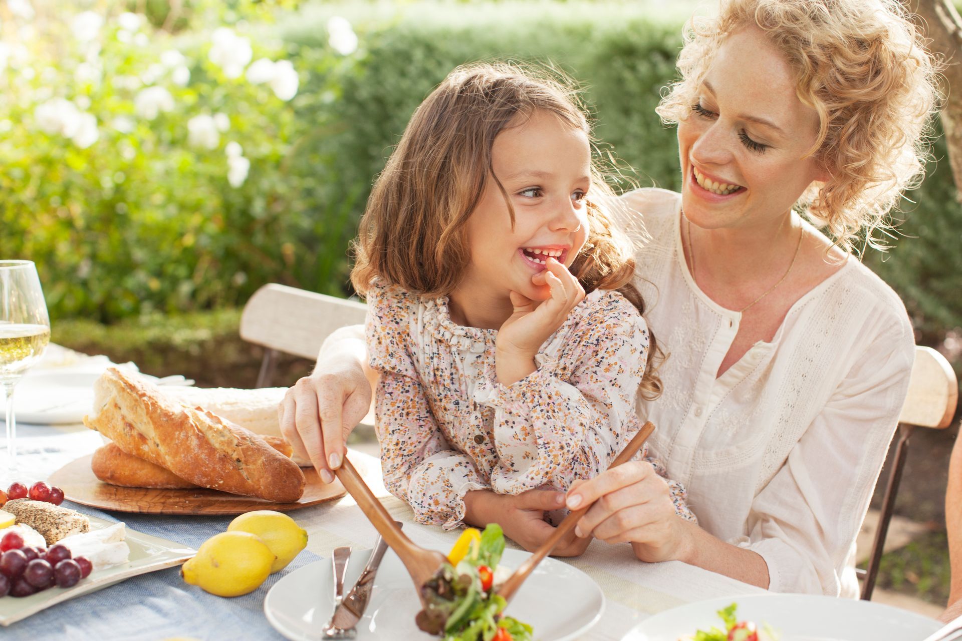 Woman and child smiling, eating salad at outdoor table with bread and fruit.