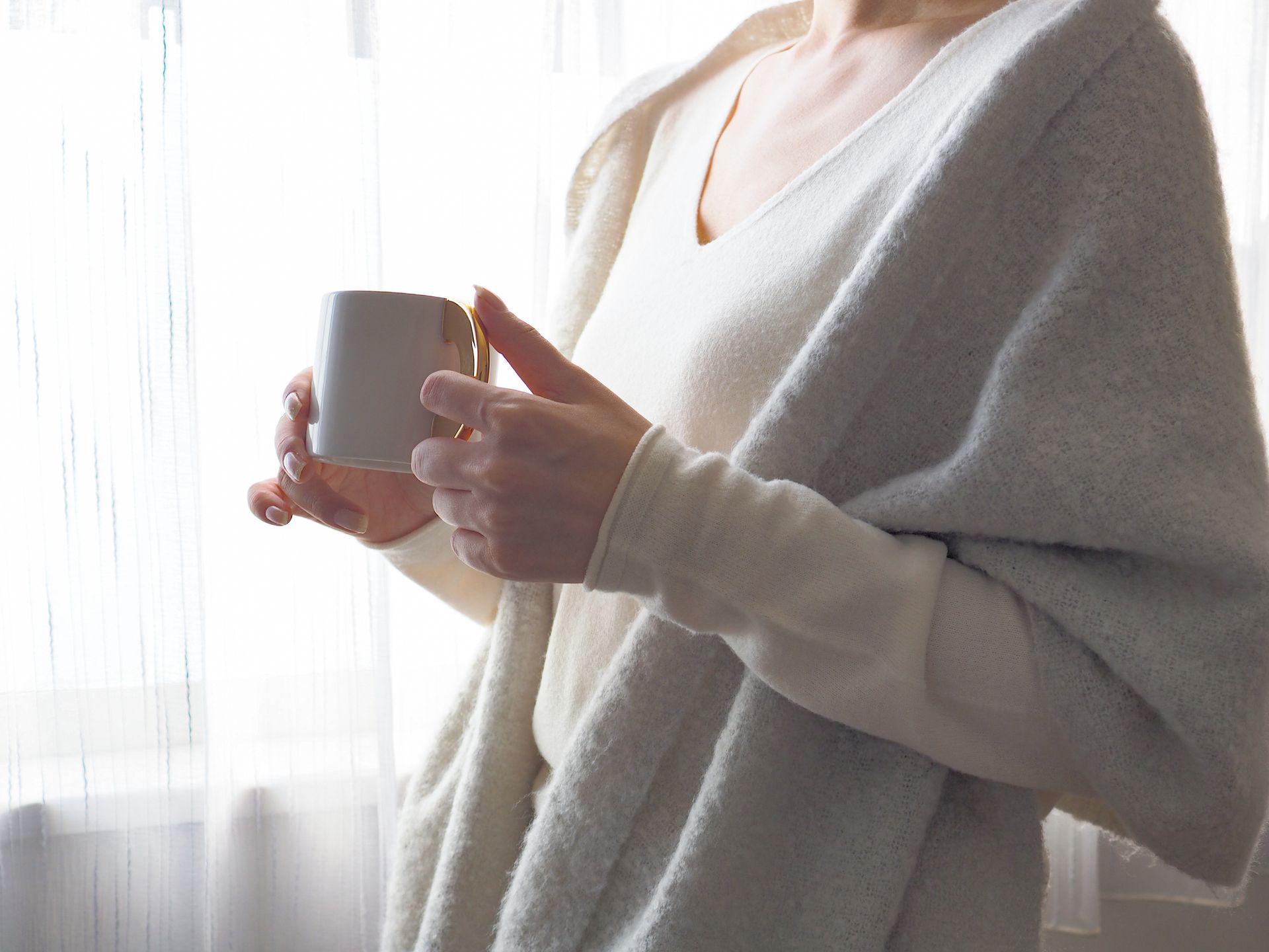 Person holding a mug, wrapped in a light-colored shawl, standing by a window.